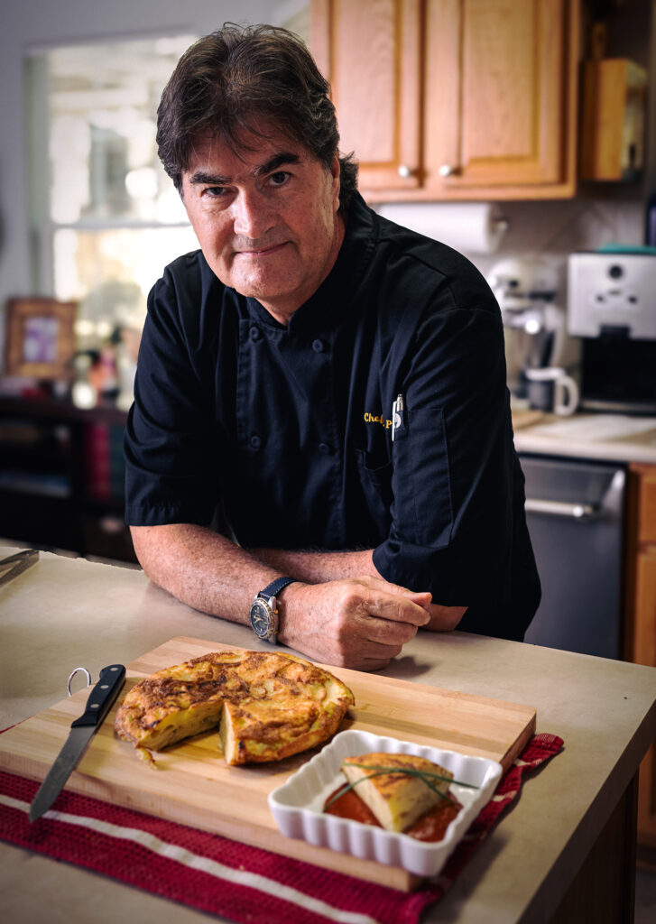 Environmental portrait of chef Eric Paz wearing a black chef's jacket in his kitchen, standing behind a freshly prepared Spanish tortilla. Photography by Javier Shirley, Austin, Texas.