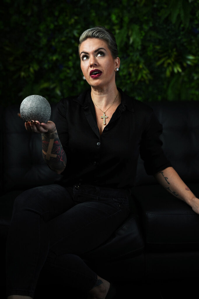 Creative editorial portrait of a short-haired woman in black, holding a silver sphere while looking upward, against a dark green background. Photography by Javier Shirley.