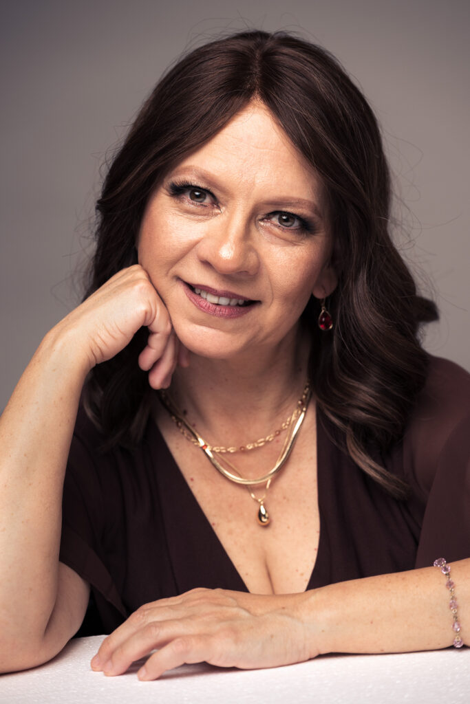 Fine art studio portrait of Olga wearing dark hair, a red blouse and gold jewelry, resting her chin on her hand against a neutral background. Photography by Austin portrait photographer Javier Shirley.