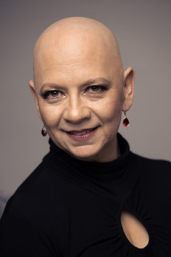 Fine art studio portrait of Olga, a bald woman wearing a black outfit and red drop earrings, looking directly at the camera against a neutral gray background. Photography by Javier Shirley.